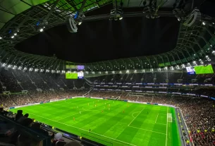 Vista interna de um estádio de futebol moderno, iluminado à noite, com arquibancadas lotadas e jogo em andamento no campo gramado.