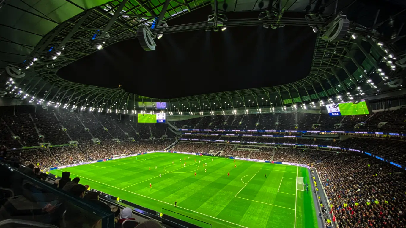 Vista interna de um estádio de futebol moderno, iluminado à noite, com arquibancadas lotadas e jogo em andamento no campo gramado.