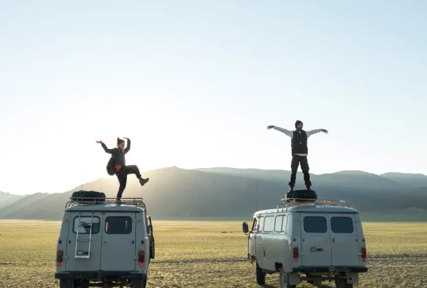 Duas pessoas em pé sobre vans em um campo aberto ao pôr do sol, celebrando a liberdade de viajar.