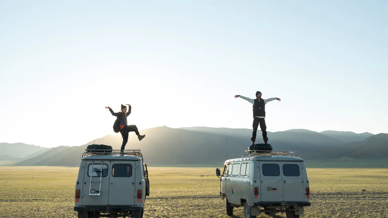 Duas pessoas em pé sobre vans em um campo aberto ao pôr do sol, celebrando a liberdade de viajar.