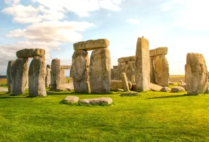 Formação de pedras monumentais de Stonehenge ao entardecer, com céu parcialmente nublado e gramado verde ao redor.