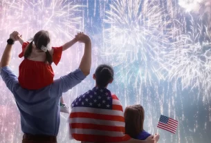 Uma família assiste a um espetáculo de fogos de artifício no 4 de Julho. Um adulto carrega uma criança no ombro, enquanto outra criança ao lado segura uma bandeira americana. Todos estão com as costas voltadas, observando os fogos coloridos iluminando o céu noturno, com a bandeira americana enrolada no adulto.