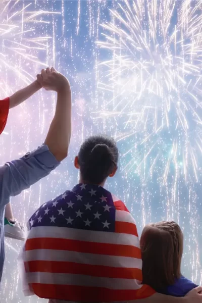 Uma família assiste a um espetáculo de fogos de artifício no 4 de Julho. Um adulto carrega uma criança no ombro, enquanto outra criança ao lado segura uma bandeira americana. Todos estão com as costas voltadas, observando os fogos coloridos iluminando o céu noturno, com a bandeira americana enrolada no adulto.