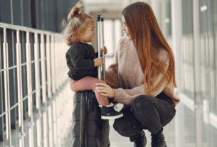 Mãe sorrindo para sua filha pequena sentada em uma mala no corredor de um aeroporto.