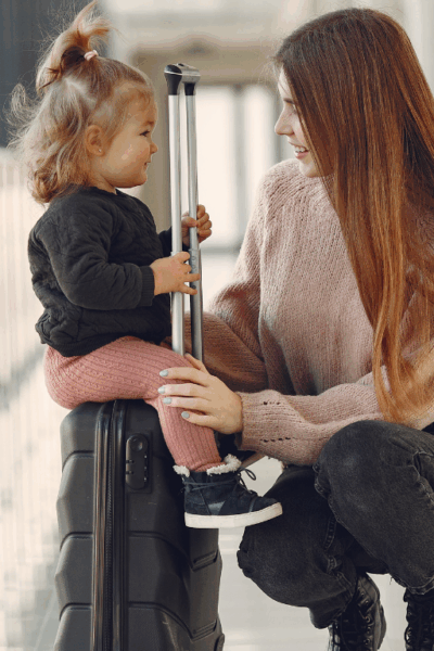 Mãe sorrindo para sua filha pequena sentada em uma mala no corredor de um aeroporto.