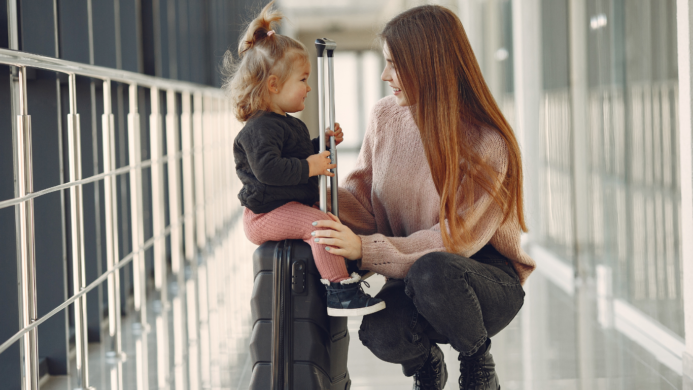 Mãe sorrindo para sua filha pequena sentada em uma mala no corredor de um aeroporto.