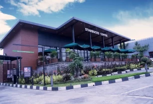 Fachada moderna de uma loja Starbucks com área externa coberta por ombrelones verdes, cercada por paisagismo tropical e ambiente urbano ao fundo.