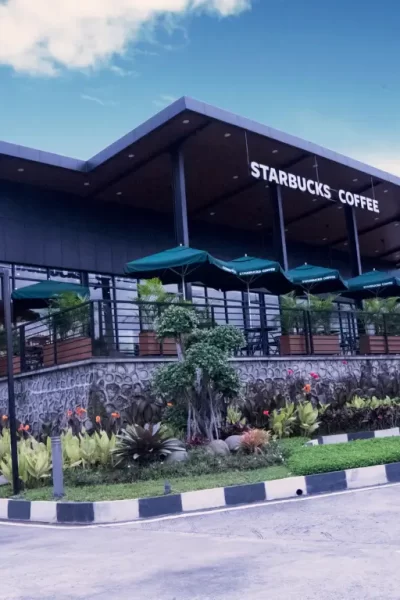 Fachada moderna de uma loja Starbucks com área externa coberta por ombrelones verdes, cercada por paisagismo tropical e ambiente urbano ao fundo.