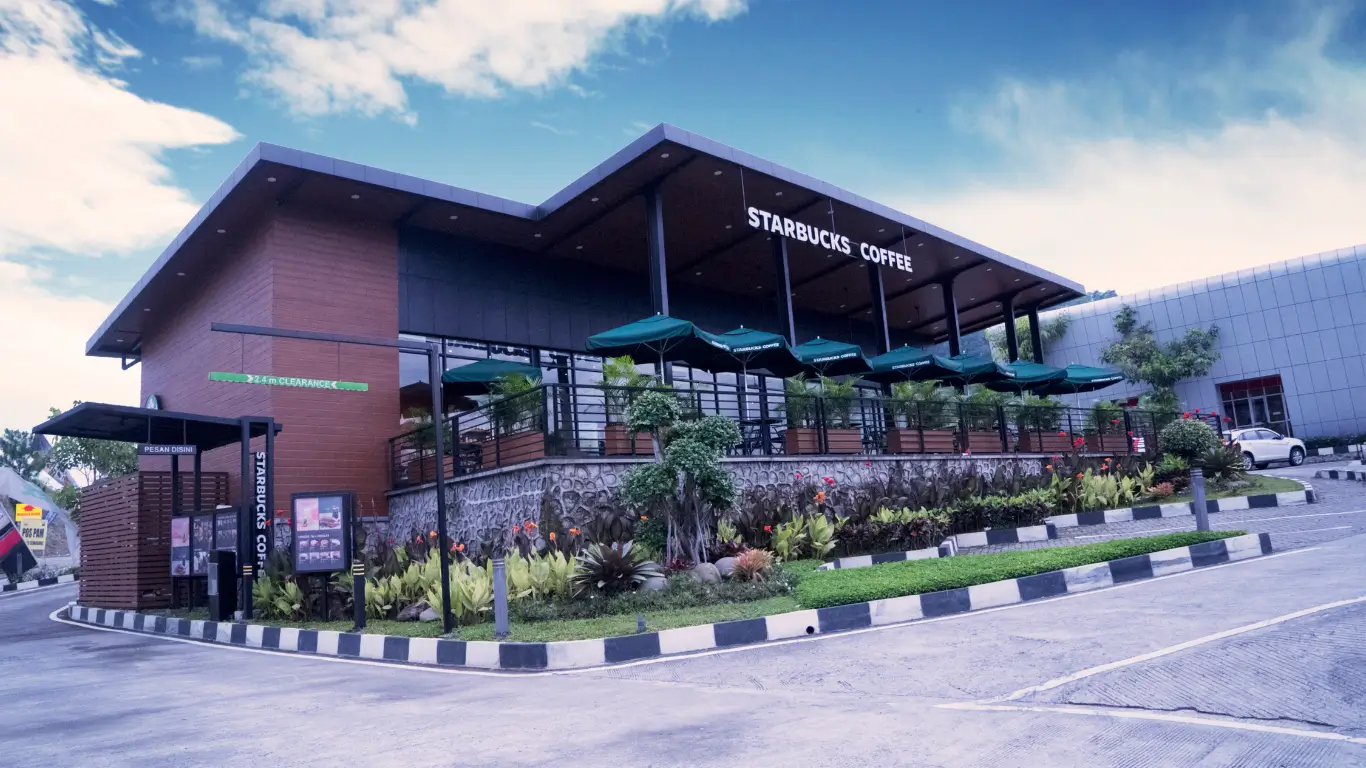 Fachada moderna de uma loja Starbucks com área externa coberta por ombrelones verdes, cercada por paisagismo tropical e ambiente urbano ao fundo.
