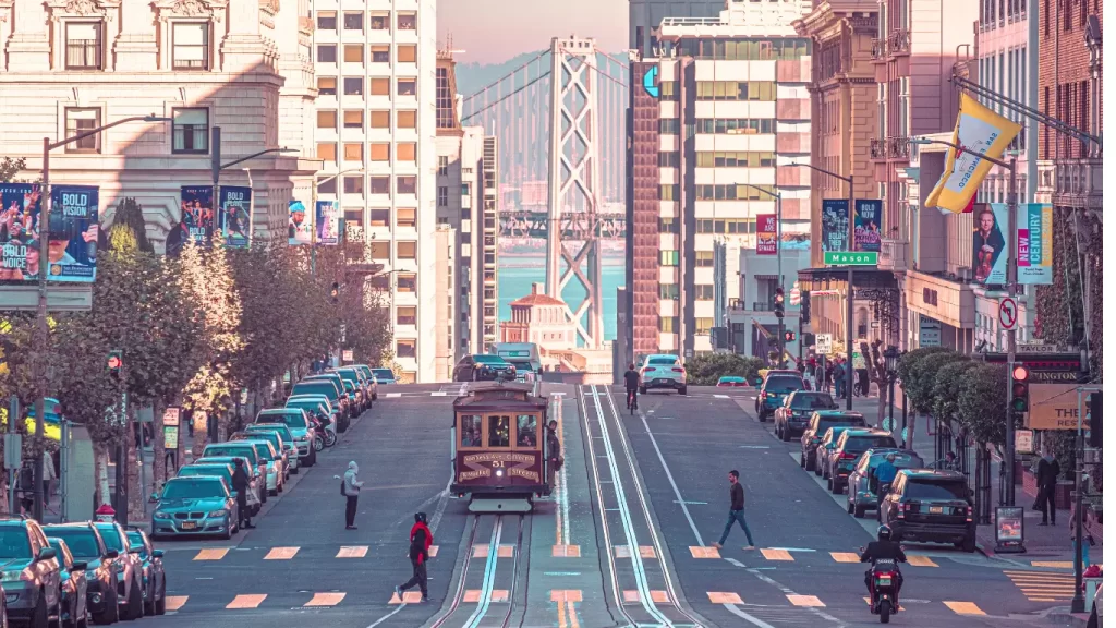 Rua urbana com bondinho, carros estacionados e arranha-céus, ao estilo urbano de grande cidade.
