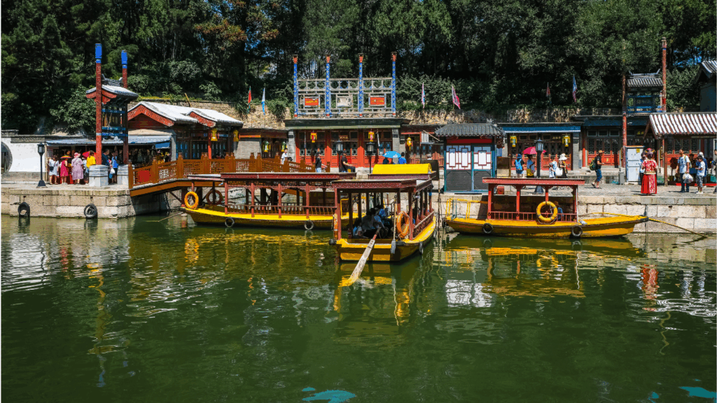 Barcos amarrados e navegando em um lago cercado por construções de madeira coloridas e vegetação, com várias pessoas ao fundo em um píer turístico.