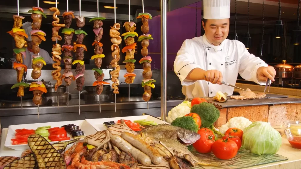 "Chef preparando churrasco gourmet com espetos de carnes, legumes frescos e frutos do mar em uma estação de grelhados."