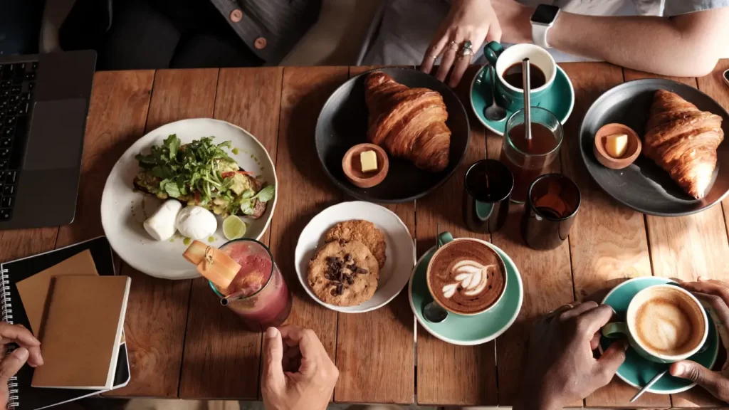 "Café da manhã gourmet com croissants, cappuccino, salada fresca, cookies e suco natural sobre mesa de madeira."