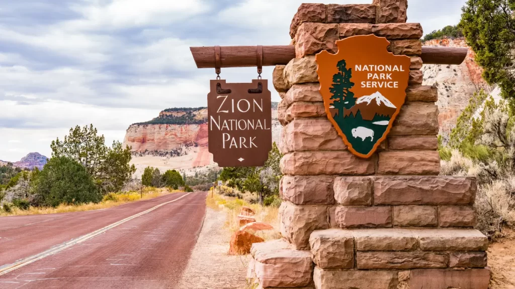 Sinal de entrada do Zion National Park ao lado de uma estrada, com rochas vermelhas ao fundo e o emblema do National Park Service.