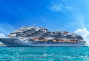 Uma grande embarcação de cruzeiro branca flutua em águas cristalinas sob um céu azul claro com algumas nuvens esparsas. A nave possui vários decks com janelas e áreas de lazer, incluindo uma piscina no topo.