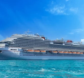 Uma grande embarcação de cruzeiro branca flutua em águas cristalinas sob um céu azul claro com algumas nuvens esparsas. A nave possui vários decks com janelas e áreas de lazer, incluindo uma piscina no topo.