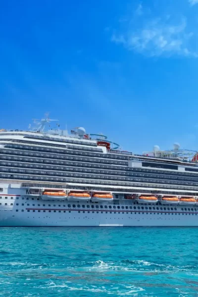 Uma grande embarcação de cruzeiro branca flutua em águas cristalinas sob um céu azul claro com algumas nuvens esparsas. A nave possui vários decks com janelas e áreas de lazer, incluindo uma piscina no topo.