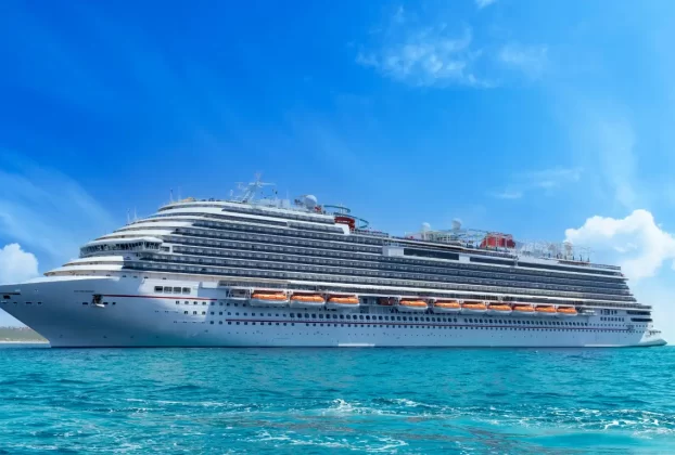 Uma grande embarcação de cruzeiro branca flutua em águas cristalinas sob um céu azul claro com algumas nuvens esparsas. A nave possui vários decks com janelas e áreas de lazer, incluindo uma piscina no topo.
