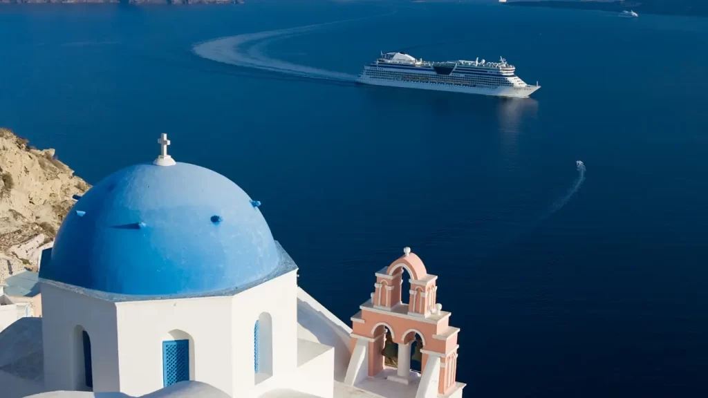 Uma igreja grega tradicional com cúpula azul e campanário rosa ao lado de um penhasco, com um grande navio de cruzeiro navegando em um mar azul profundo ao fundo.
